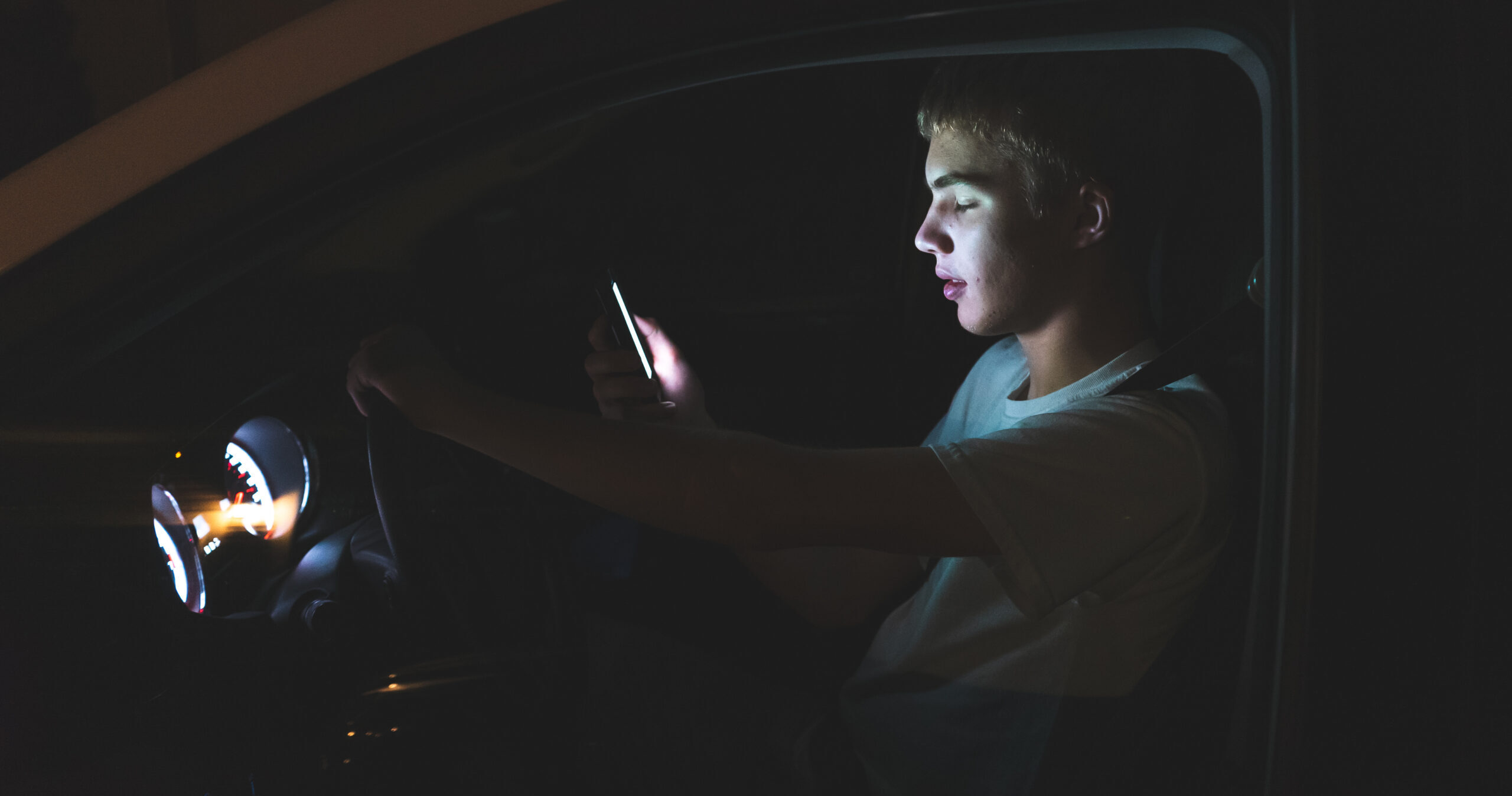 Distracted teenager driving a car with his cell phone in his hand. The light from the screen of the phone is illuminating his face.